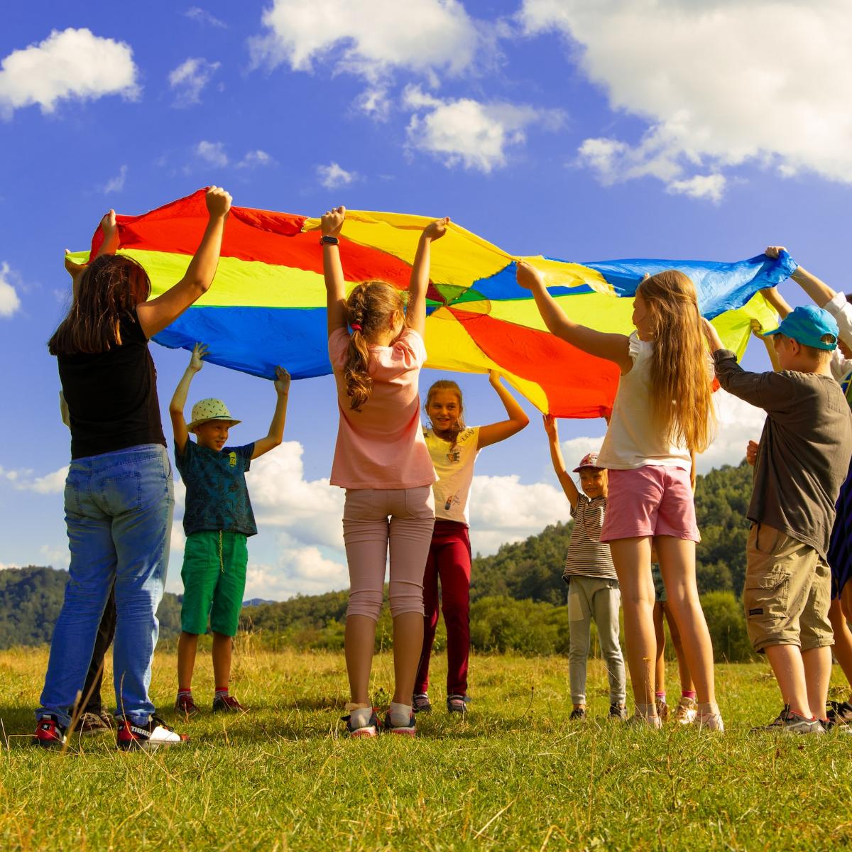 Eine Gruppe Kinder hält ein buntes 'Schwungtuch' in die Luft. Sie stehen auf einer Wiese. Der Himmel ist blau-weiß bewölkt