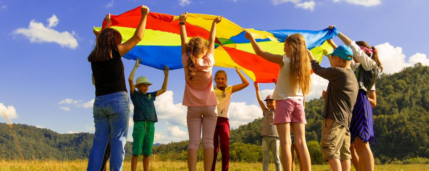 Eine Gruppe Kinder hält ein buntes 'Schwungtuch' in die Luft. Sie stehen auf einer Wiese. Der Himmel ist blau-weiß bewölkt