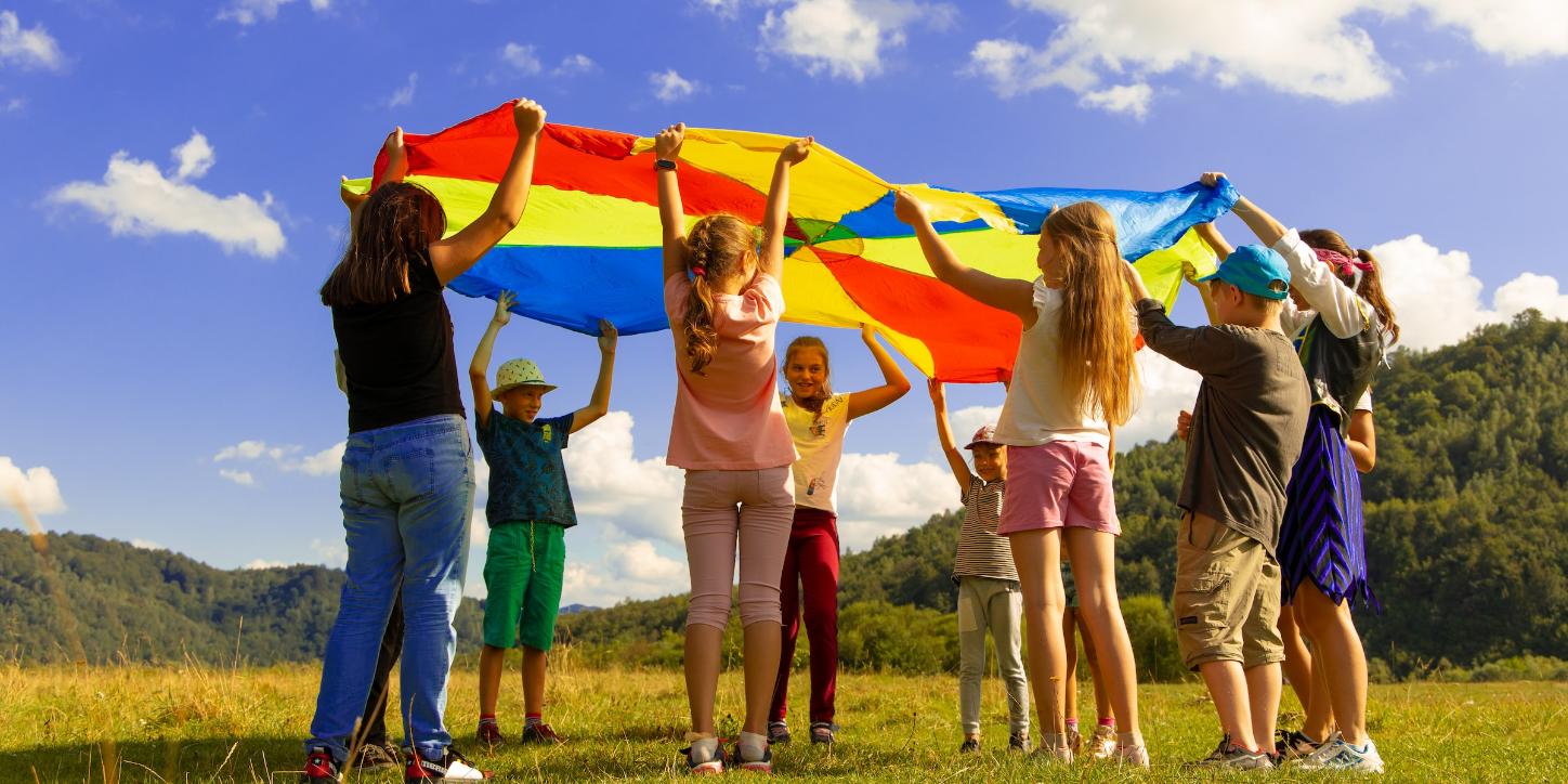 Eine Gruppe Kinder hält ein buntes 'Schwungtuch' in die Luft. Sie stehen auf einer Wiese. Der Himmel ist blau-weiß bewölkt