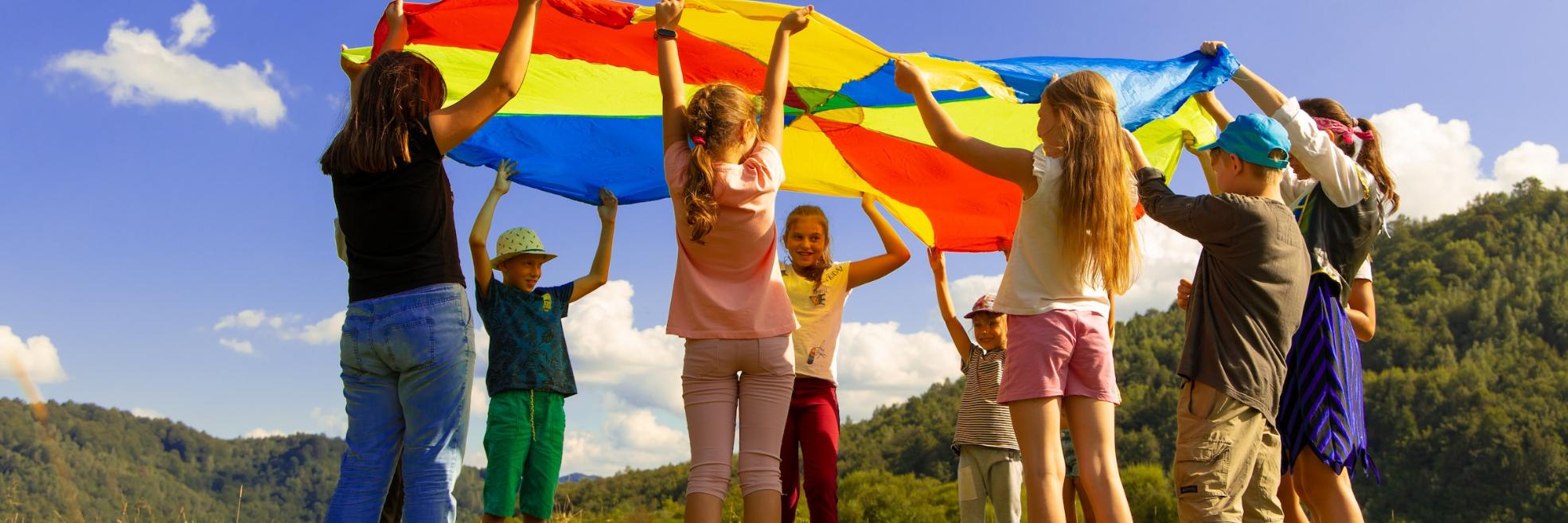 Eine Gruppe Kinder hält ein buntes 'Schwungtuch' in die Luft. Sie stehen auf einer Wiese. Der Himmel ist blau-weiß bewölkt